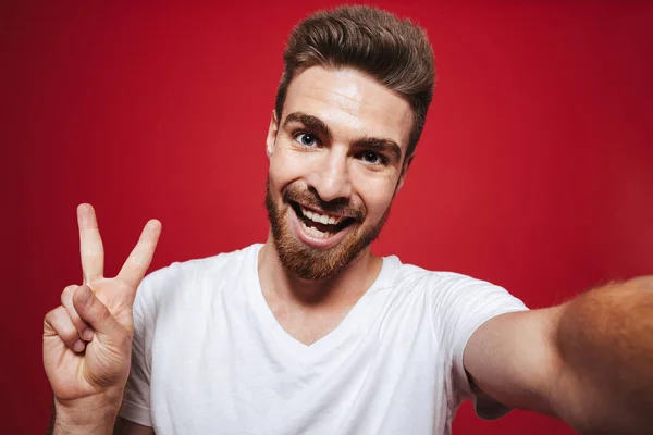 赤い背景に分離された Selfie をしながら平和のジェスチャーを示す陽気な若いひげを生やした男の肖像 — ストック写真