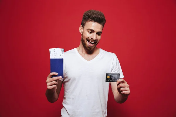 一个开朗的年轻胡子男子的肖像持有护照与门票和显示塑料信用卡隔离红色背景 — 图库照片