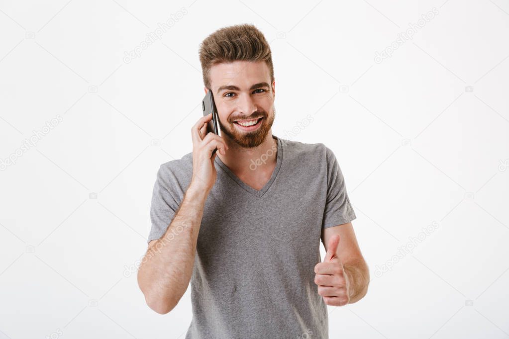 Portrait of a happy young bearded man talking on mobile phone and showing thumbs up isolated over white background