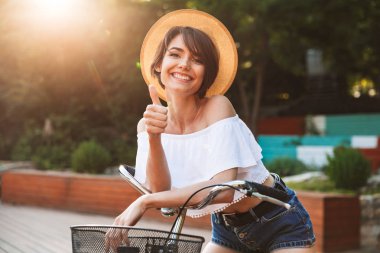 Parkta Bisiklete binmek ve başparmak gösterilen yazlık giysiler içindeki genç kız gülümseyerek