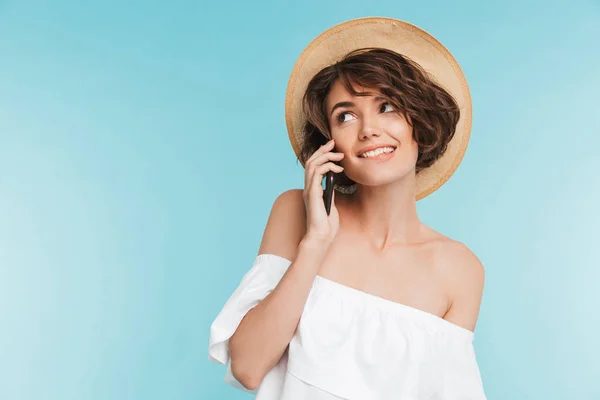 一个快乐的年轻女子在夏季帽子的肖像谈论在蓝色背景下的手机隔离 — 图库照片