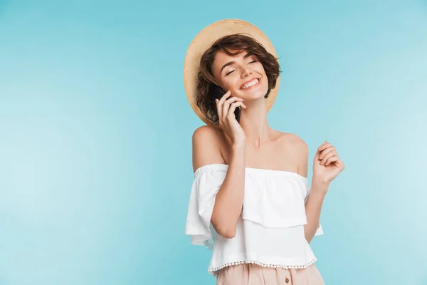 一个微笑的年轻女子在夏季帽子的肖像谈论在蓝色背景下的手机隔离 — 图库照片