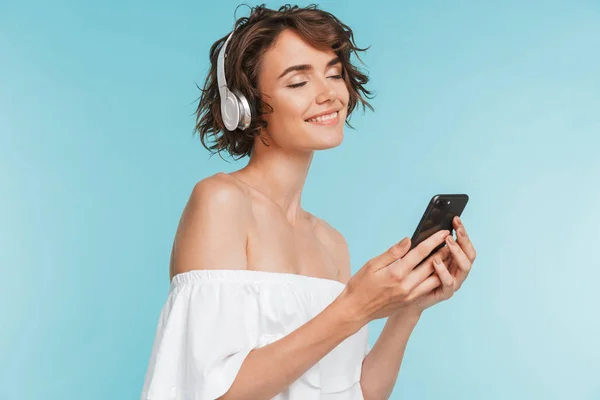 ヘッドフォンと青い背景に分離された携帯電話で音楽を聴く笑顔の若い女性の肖像画 — ストック写真
