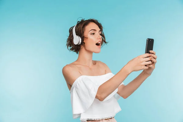 一个漂亮的年轻女子的肖像 听音乐与耳机 并采取自拍与手机隔离在蓝色背景 — 图库照片