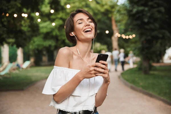 イヤホンと公園で立ちながら携帯電話で音楽を聴く幸せな若い女の子 — ストック写真