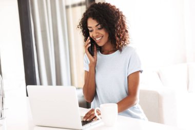 Fincan kahve ile evde dizüstü bilgisayar kullanırken cep telefonda konuşurken gülümseyen Afrika kadın