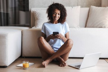 Bardak portakal suyu ve defter ile evde bir kanepede otururken cep telefonuyla gülümseyen Afrika kadın