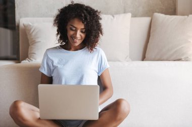 Evde bir kanepede otururken dizüstü bilgisayar kullanan gülümseyen Afrika kadın