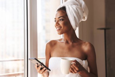 Oldukça Afrikalı kadın cep telefonuyla dururken pencerenin önünde evde bir havlu sarılı