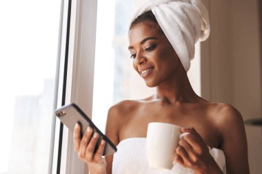 Cep telefonuyla dururken pencerenin önünde evde bir havlu sarılı güzel Afrikalı kadın