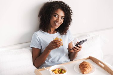 Gülümseyen genç Afrikalı kadın yatakta bir tepsi üzerinde lezzetli kahvaltı yaparken cep telefonunuzun