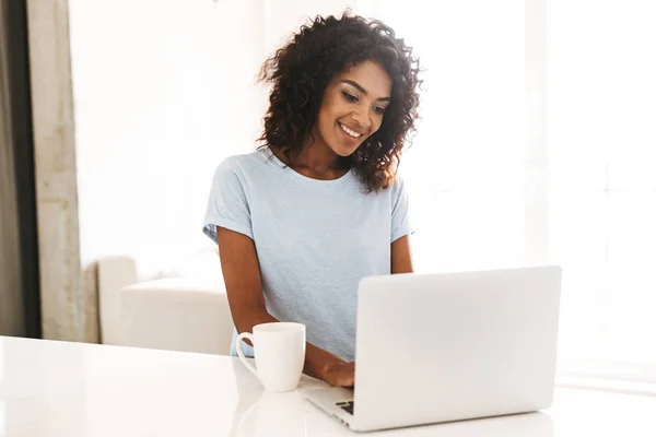坐在家里喝咖啡的非洲女人微笑着使用笔记本电脑 — 图库照片