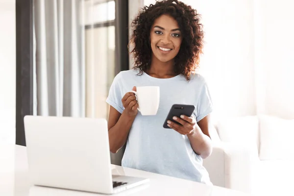 愉快的非洲妇女使用移动电话 当拿着杯子咖啡和坐与膝上型电脑在家里 — 图库照片