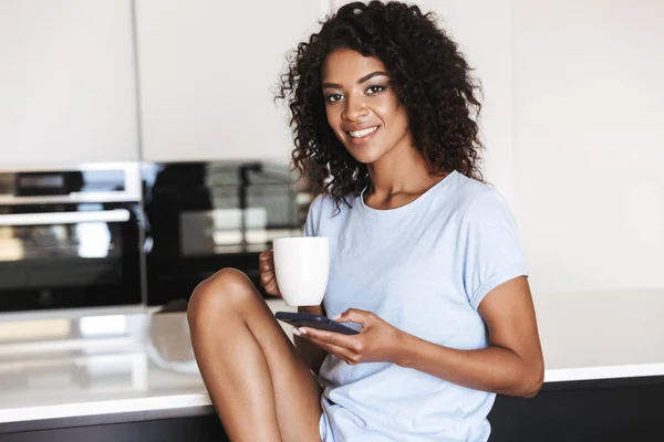 满意的非洲妇女使用手机 而坐在椅子上 在家里的一杯咖啡 — 图库照片