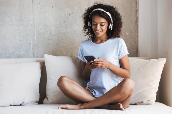 Smiling african woman in headphones using mobile phone while sitting on a couch at home