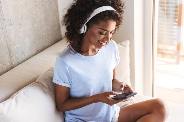 Smiling african woman in headphones using mobile phone while sitting on a couch at home