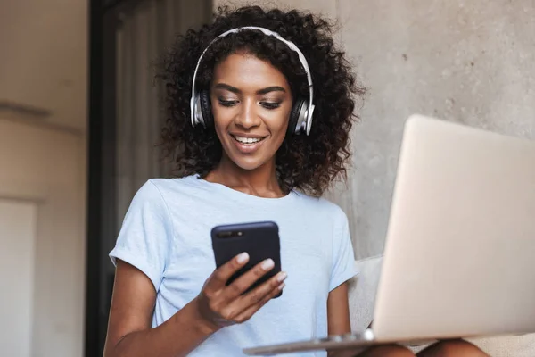 Leende Afrikanska Kvinna Hörlurar Använder Mobiltelefon Medan Sitter Med Bärbar — Stockfoto