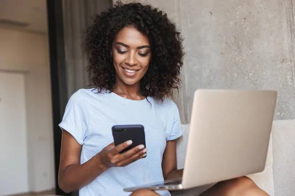 在家里的沙发上坐着膝上型电脑的非洲女人微笑着使用手机 — 图库照片