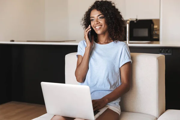 笑顔でラップトップ コンピューター 自宅でソファに座って携帯電話で話しているアフリカの女性 — ストック写真