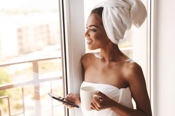Усміхнена Африканка Загорнута Рушник Використовує Мобільний Телефон Стоячи Біля Вікна — стокове фото