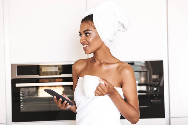 Happy african woman wrapped in a towel using mobile phone and hoding cup of coffee while standing at the kitchen