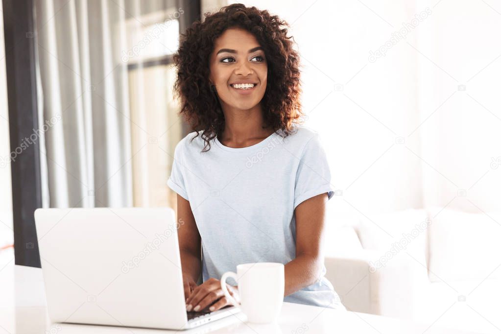 Cheerful african woman using laptop computer while sitting at home with cup of coffee