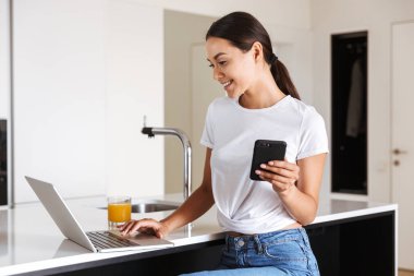 Cep telefonu ve bardak portakal suyu olan bir mutfak üzerinde otururken dizüstü kullanarak çekici genç Asyalı kadın