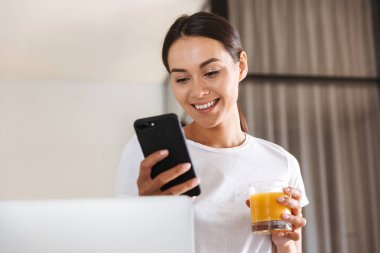 Çekici genç Asyalı kadın cep telefonu duran bir mutfak bardak suyu ile kullanma