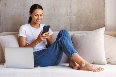 Evde dizüstü bilgisayarla koltukta otururken cep telefonu kullanan gülümseyen Asyalı genç kadın.