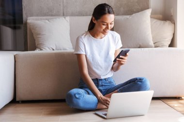 Mutlu Genç Asyalı kadın dizüstü bilgisayar ile evde bir kanepede otururken cep telefonunuzun