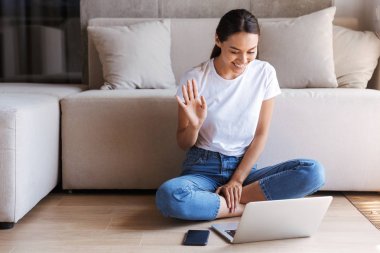 Dizüstü bilgisayar ile bir katta otururken bir videocall sahip neşeli Genç Asyalı kadın