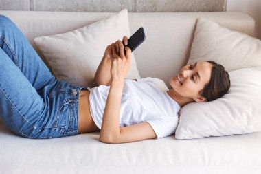 Neşeli Genç Asyalı kadın evde bir kanepede döşenmesi sırasında cep telefonuyla