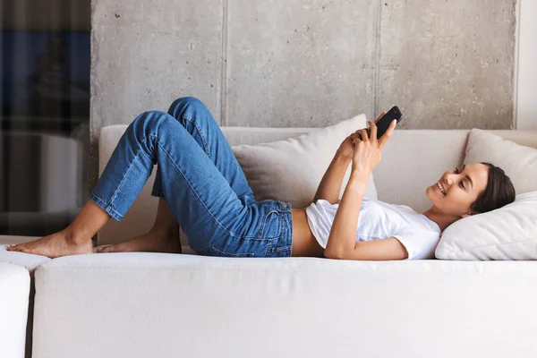 笑顔の若いアジア女性 自宅でソファの上に敷設しながら携帯電話を使用して — ストック写真
