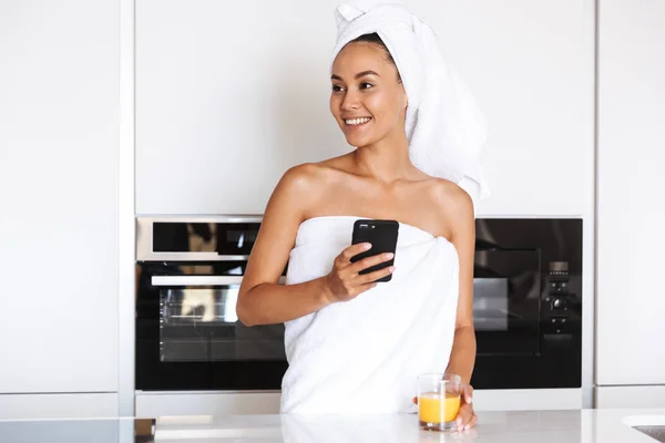 年轻的亚洲妇女 用手机裹在淋浴毛巾上 站在厨房里捧着一杯果汁 — 图库照片