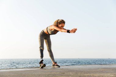 Full length of focused disabled athlete woman with prosthetic leg doing stretching exercises while standing at the beach clipart