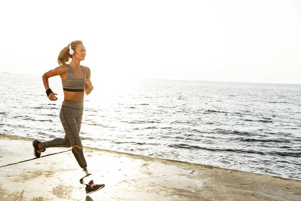 Image of young amazing disabled sports woman running on the beach outdoors listening music with headphones.