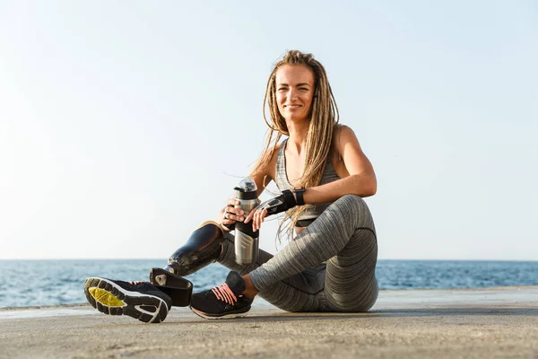 Smiling disabled athlete woman with prosthetic leg holding water bottle while sitting at the beach