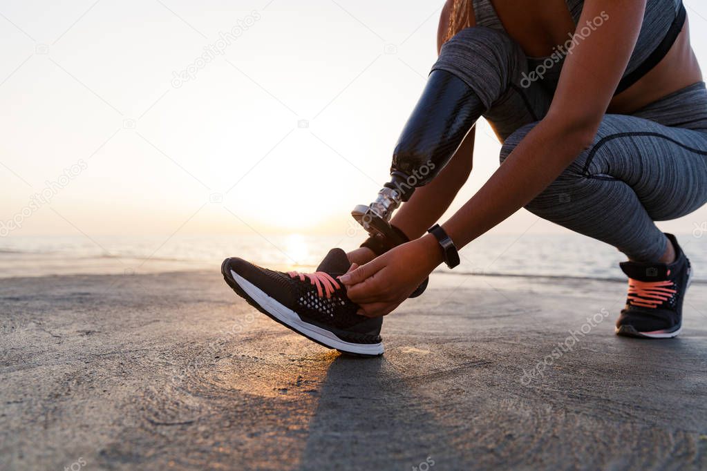Close up of disabled athlete woman with prosthetic leg tying her shoelace outdoor at the beach