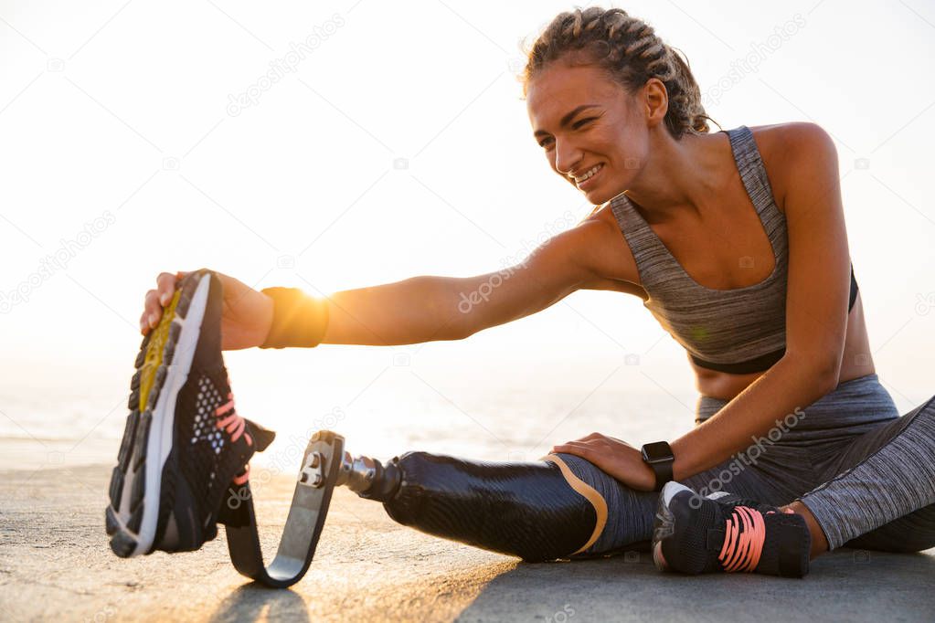 Cropped image of smiling disabled athlete woman with prosthetic leg doing stretching exercises while sitting at the beach