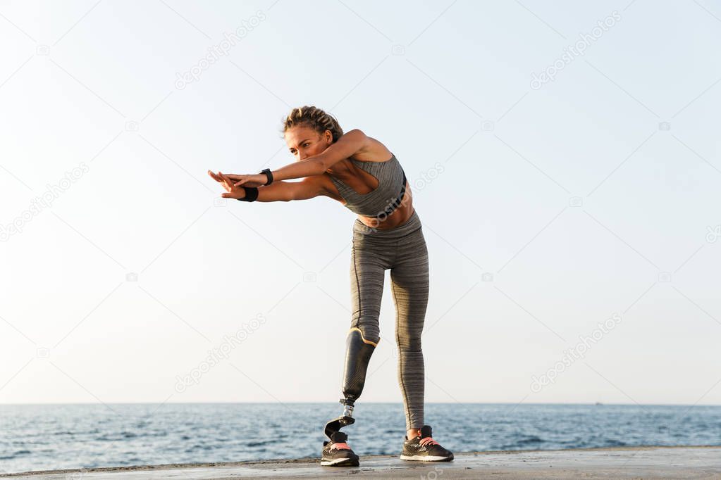 Full length of motivated disabled athlete woman with prosthetic leg doing stretching exercises while standing at the beach
