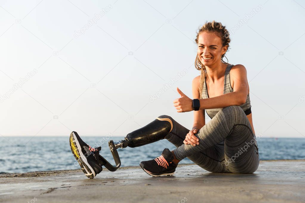 Smiling disabled athlete woman with prosthetic leg showing thumbs up while sitting at the beach
