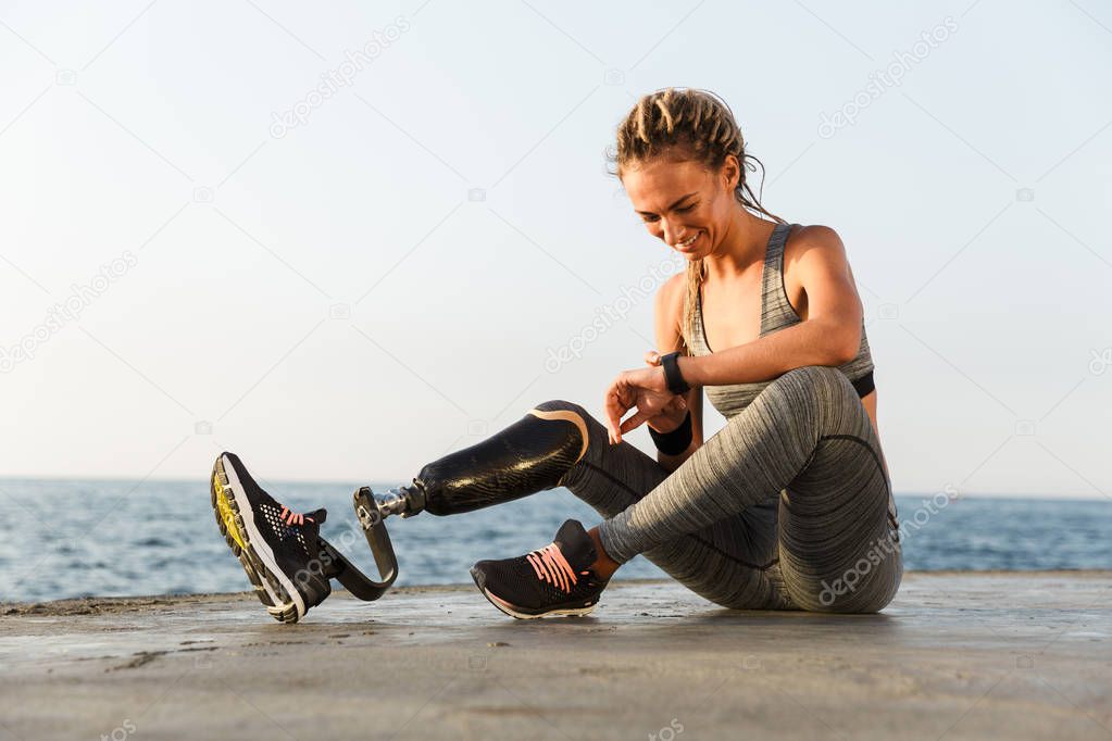 Smiling disabled athlete woman with prosthetic leg checking smart watch while sitting at the beach