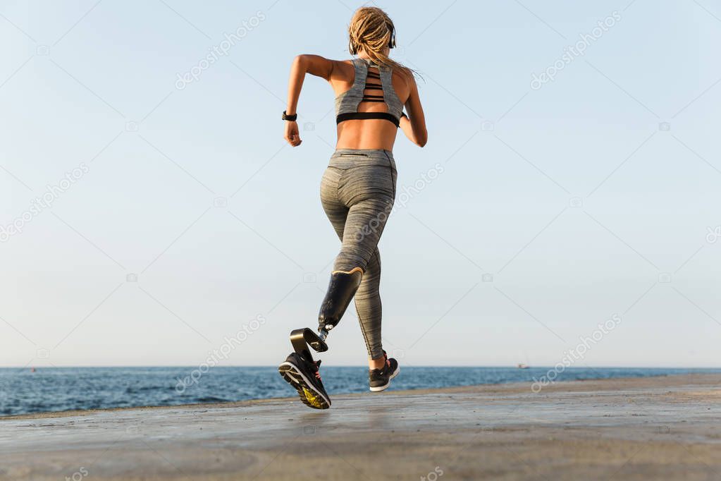 Back view of young amazing disabled sports woman running on the beach outdoors listening music with headphones.