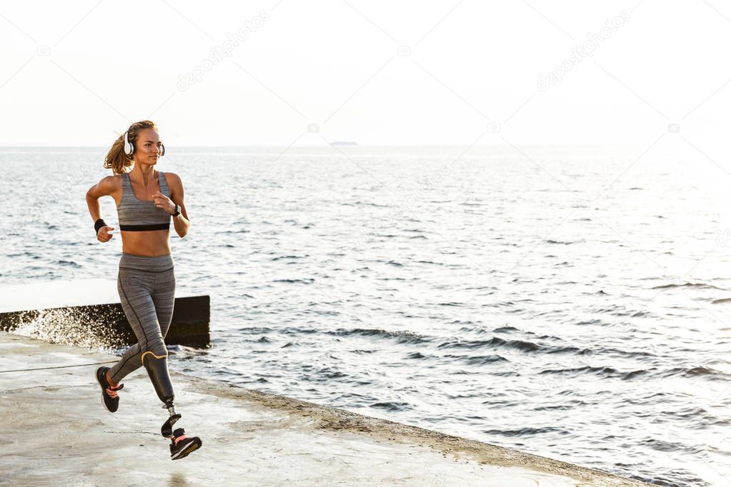 Image of young amazing disabled sports woman running on the beach outdoors listening music with headphones.
