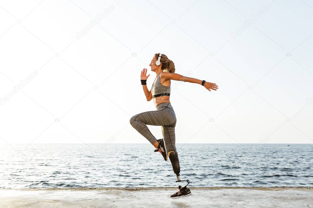 Photo of young strong disabled sports woman running on the beach outdoors listening music with headphones.