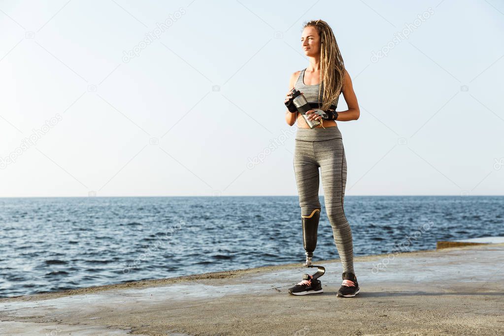 Full length of smiling disabled athlete woman with prosthetic leg standing at the beach and drinking water from a bottle