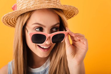 Uzak izole sarı arka plan üzerinde neşeli genç sarışın bir kadın yaz şapka ve güneş gözlüğü seyir portresi kapatın