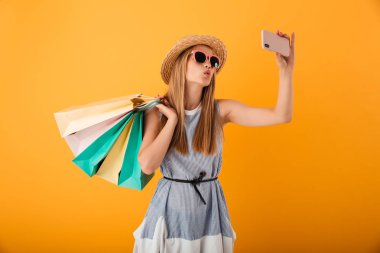 Güzel bir sarışın genç kadın yaz şapka ve güneş gözlüğü alışveriş torbaları üzerinde sarı arka plan izole tutarken bir selfie alarak portresi