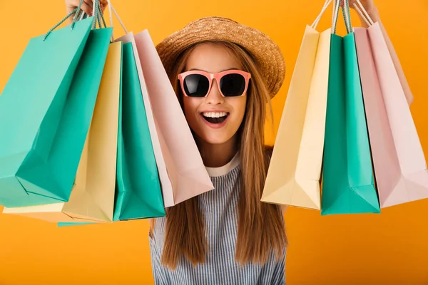 一个满意的年轻金发女郎的肖像在夏季帽子和太阳镜持有购物袋 隔离在黄色背景 — 图库照片