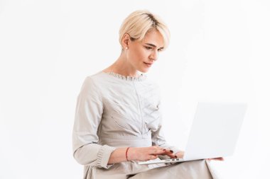 Elbise oturma ve Studio beyaz arka plan üzerinde izole gümüş dizüstü bilgisayar kullanarak kısa sarı saçları zarif orta yaşlı kadınla